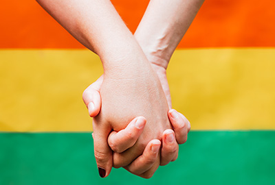 Im Hintergrund Ausschnitt einer Regenbogenflagge - im Vordergrund händchenhaltende Frauen. Sichtbar sind Unterarm und Hände