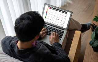 Junger Mann von hinten mit Laptop auf den Beinen gibt Daten in einen Onlinekalender ein